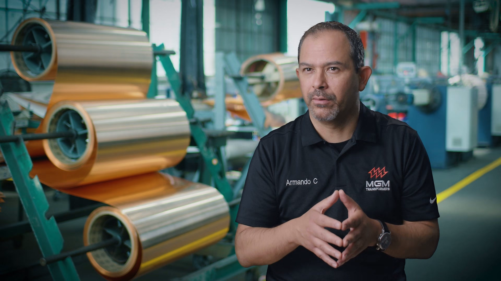Armando C of MGM Transformers speaking on the factory floor, bright copper rolls racked behind him