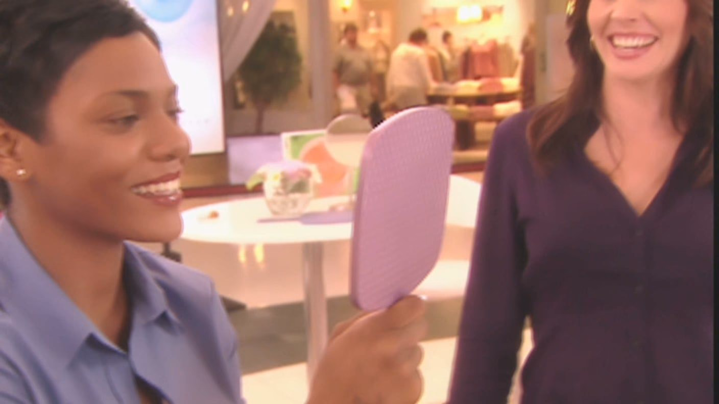 A woman smiling as she looks into a hand mirror at the Sheer Cover counter, the mall behind her