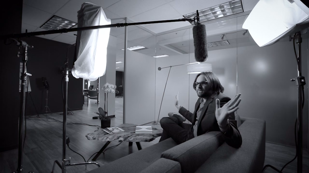 Black and white behind the scenes frame: the subject on a couch mid-gesture, a boom overhead and lighting stands either side